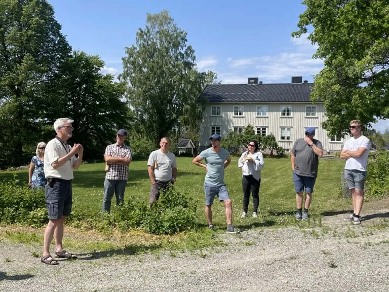 Seks personer som står i et grønt landskap med en hvit bygning i bakgrunnen.